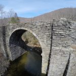 Fisheye image of old stone bridge over a river, with trees and mountains in the background.