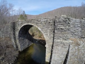 Fisheye image of old stone bridge over a river, with trees and mountains in the background.