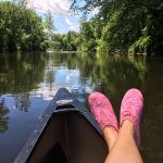 Crossed human legs with pink shoes up against the bow of a canoe in foreground, with river and green trees in background