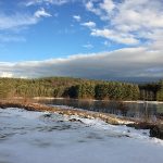 Snowy ground in foreground, water, green trees, blue sky with clouds in background