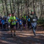 A large group of people running on the trail at the Chesterfield Gorge, wearing race bibs and dressed warmly. Sunlight shining through trees in the background.