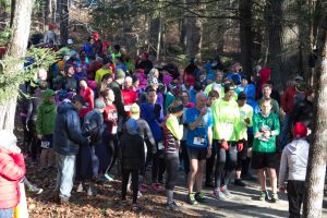 Crowd of runners dressed warmly, standing on a trail with trees in background