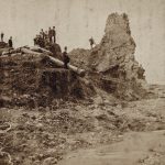 19th-century black-and-white photo of about a dozen people standing on the stone ruins of what is left of one side of the dam.