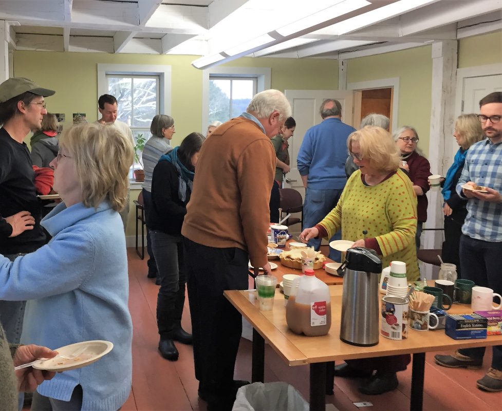 People standing around a table, helping themselves to pie and beverages, others in the room talk and look at pictures on the walls