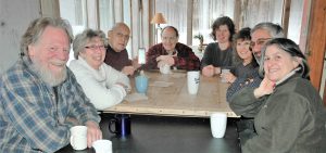 Eight people sitting around a table with mugs of hot beverages pose for a picture by smiling at the camera