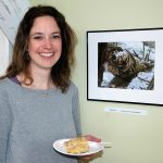 A woman is standing, holding a plate with a slice of pie. She is smiling and looking into the camera. Next to her is her winning photograph.