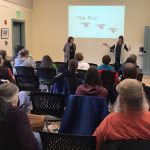 The audience is watching two women speak about their books. Behind them is a slide show with an image indicating that they are at the end of their presentation.