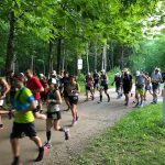 A crowd of runners take off from the starting line on a trail. Green trees are in the background.