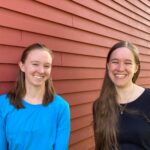 Audrey and Sarah shown from the mid-torso up, looking into the camera and smiling. Behind them is the outside wall of a red barn.