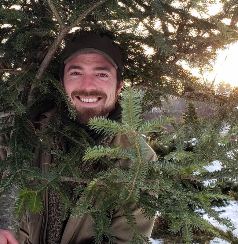 In this image, Michael, wearing a dark, brimmed hat, is mostly hidden by the branches of an evergreen tree, but his smiling face is visible, looking directly into the camera.