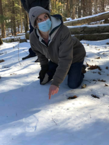 Cesi kneels in the snow to point out an animal track while looking up at a member of the group outside of the frame.