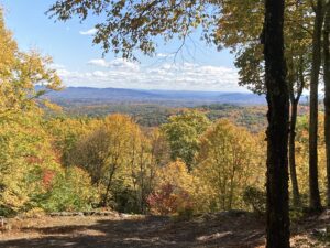 Framed by fall-colored trees, the blueish hills of the Holyoke Range are seen in the middle distance from the top of a hill.