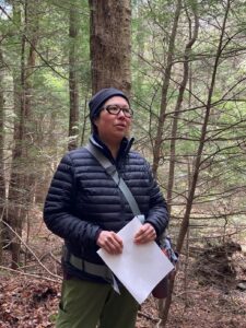 Photograph depicts JuPong Lin, holding a piece of paper, standing in the woods next to the pond and speaking.