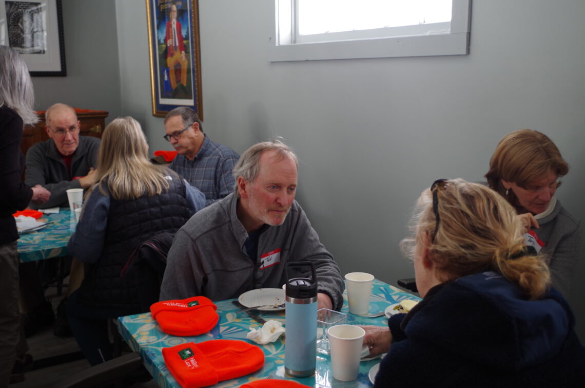 A photo of three people seated around a square table. Two seated across from each other are having a conversation, one person is listening intently to the other speaking. In front of them on the table are plates with food on them and cups.