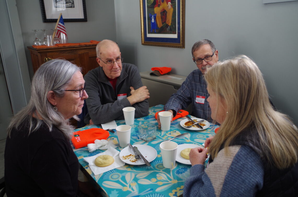 A photo of four people seated around a square table, having a conversation. In front of them  on the table are plates with food on them and cups.