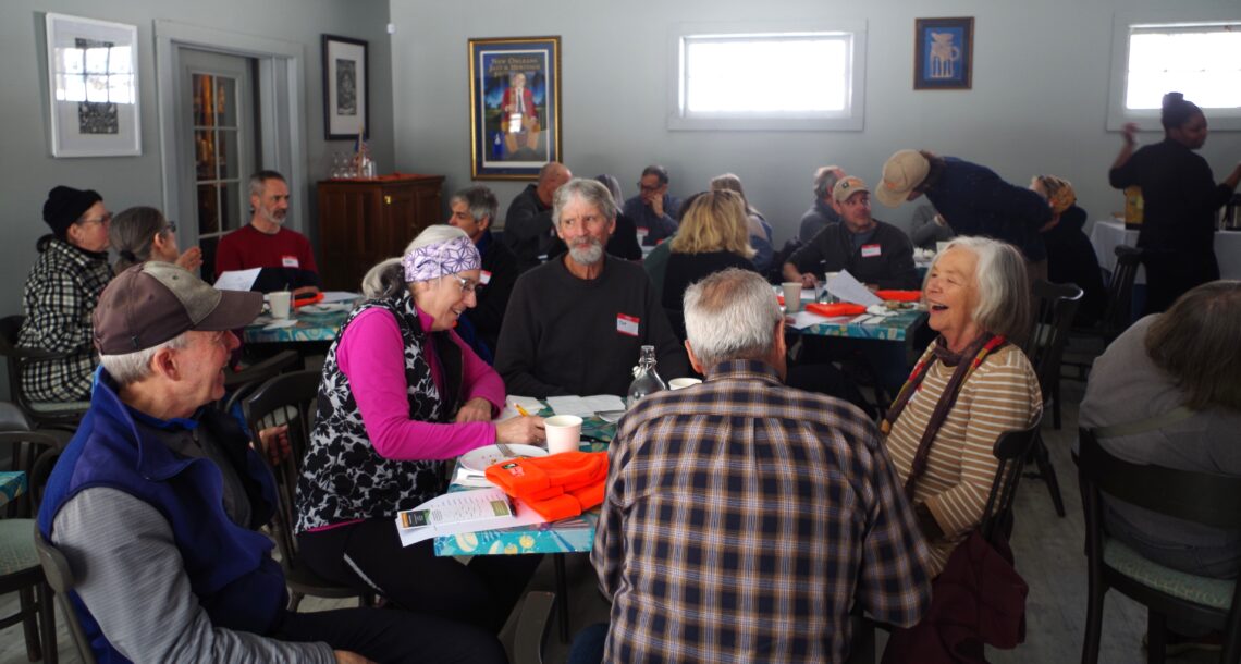 A photo of a room full of over twenty people, seated around square tables and having conversations. In the foreground is a table with five laughing people seated around it.