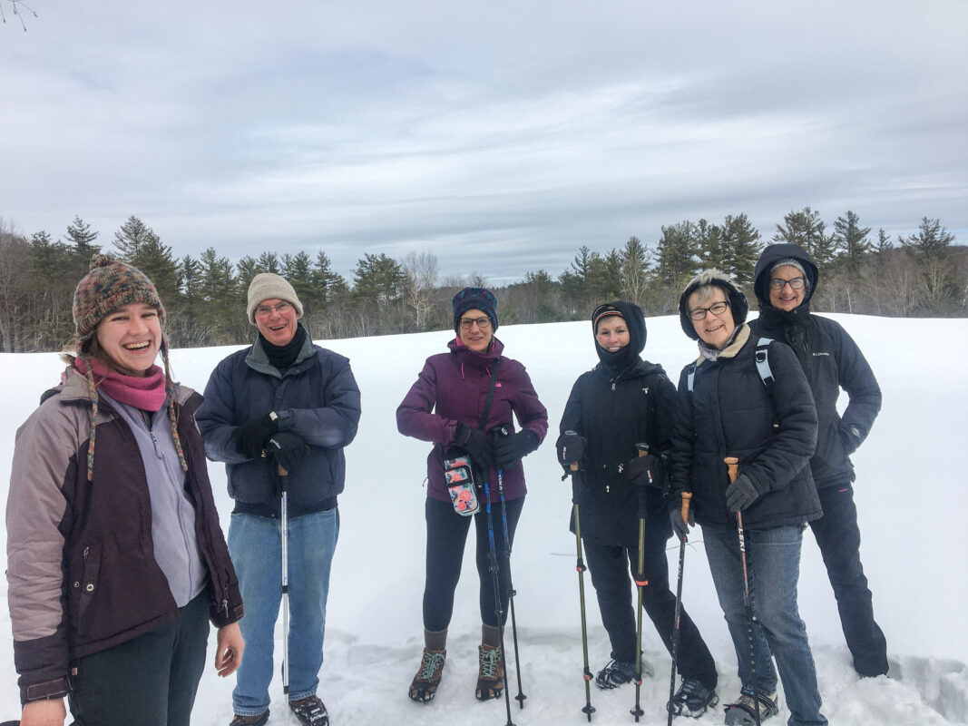 A photo of six people stand in a snow-covered field. They are dressed for winter and wearing ice-grippers on their  feet and carrying poles. They are all looking into the camera and smiling.