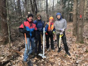 Bundled up against the cold, four people stand shoulder to shoulder in the woods, smiling into the camera and holding saws and loppers and other tools for trail work. A gray sky is visible through bare trees and a leaf covered forest floor in the background. 