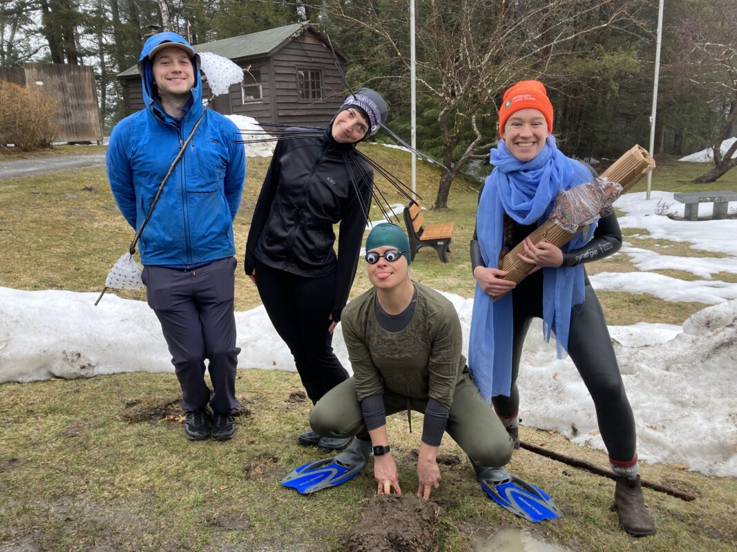 Photo of Tim, Katie, Sally, and Sarah posing for the camera. From left is: Tim wearing rain gear and a stick festooned with egg masses made out of bubble wrap. Katie dressed head to toe in black with long antennae leaning sharply over to one side like she's wiggling around. Sally, crouching in a frog pose, is dressed head to toe in green. She is wearing swim goggles with large googly eyes stuck on, and blue flippers. Sarah is holding a log with a turtle on it, both made out of cardboard, and she is wearing blue fabric draped around her neck and a blaze orange HLT hat.