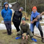 Photo of Tim, Katie, Sally, and Sarah posing for the camera. From left is: Tim wearing rain gear and a stick festooned with egg masses made out of bubble wrap. Katie dressed head to toe in black with long antennae leaning sharply over to one side like she's wiggling around. Sally, crouching in a frog pose, is dressed head to toe in green. She is wearing swim goggles with large googly eyes stuck on, and blue flippers. Sarah is holding a log with a turtle on it, both made out of cardboard, and she is wearing blue fabric draped around her neck and a blaze orange HLT hat.