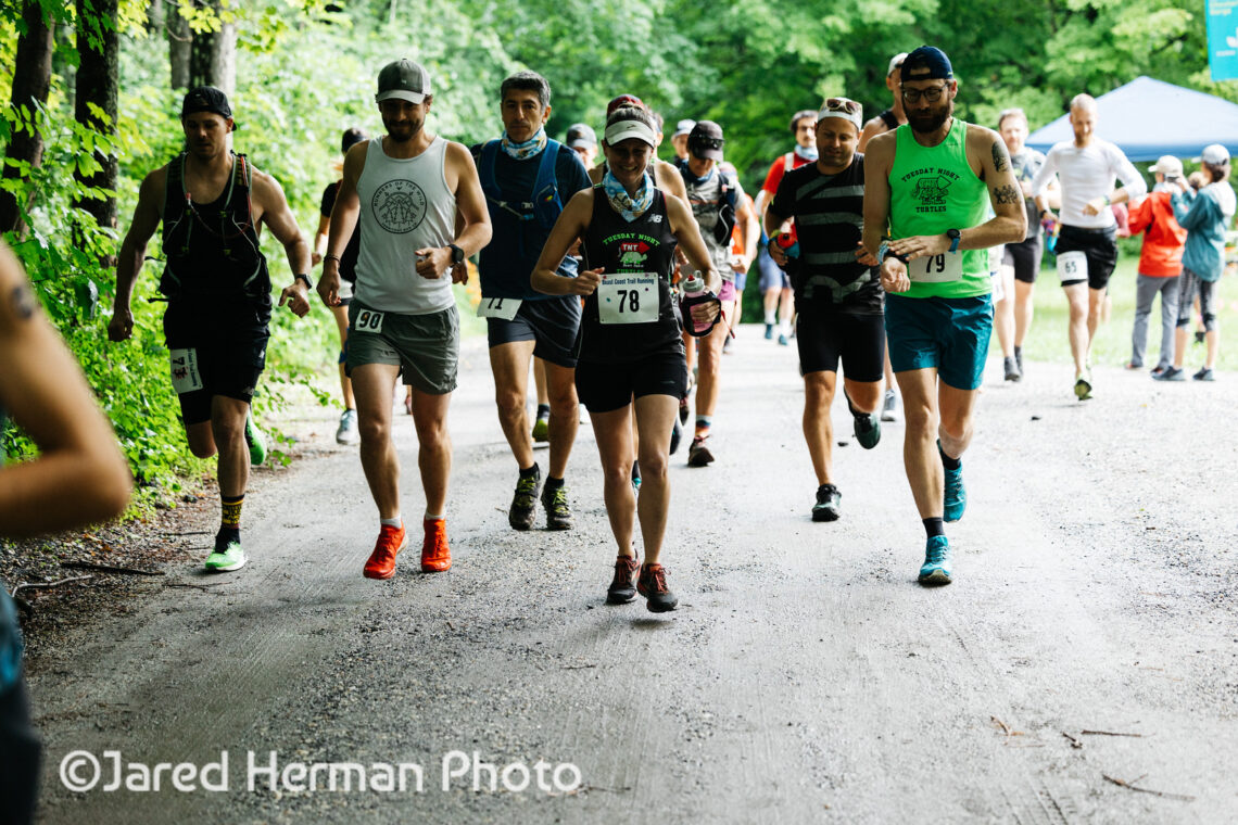 Photograph of runners in shorts, hats, and tank tops running on a dirt road. Behind them are green trees. 