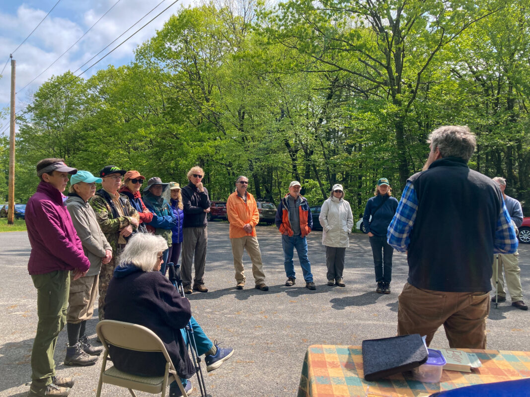 A circle of standing and seated people gathers on a quiet road with cars and trees in the background.