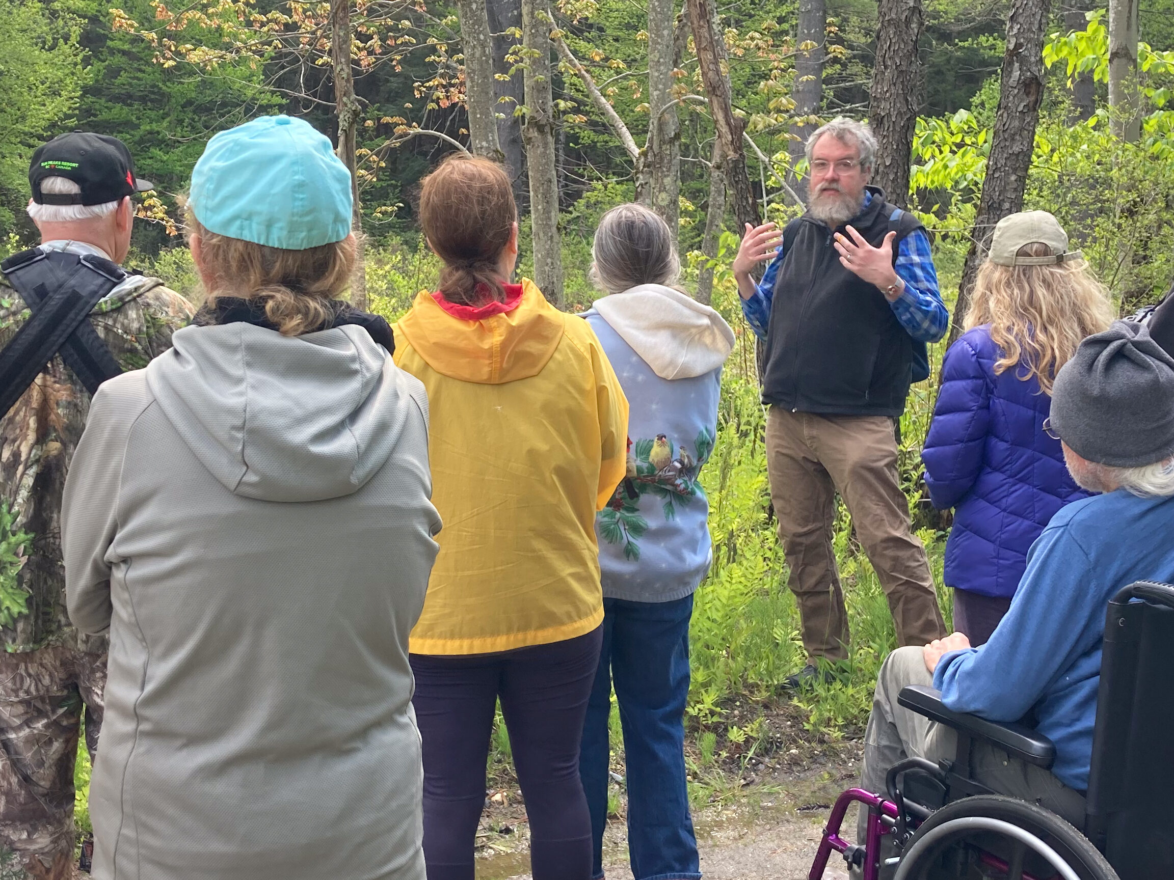 A group of standing and seated people, seen from the back, faces beaver habitat while Drew, seen facing the camera, talks about beavers.