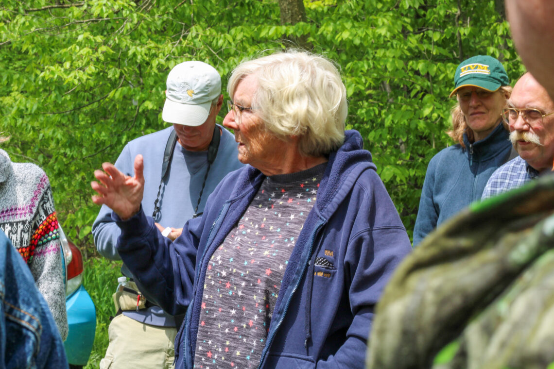 Seen from the torso up against a background of 3 people and trees, another neighbor talking to the group.