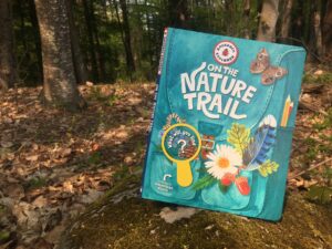 A copy of the book “Backpack Explorer: On the Nature Trail” sits on a mossy rock in a dark, leafy forest. The cover of the book is an illustration of a teal back filled with items from nature, including a blue jay feather, leaves, flowers, acorns, a caterpillar, and a magnifying glass. 