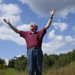 Wil Hastings, standing in a field, smiles and throws his arms joyously into the air.