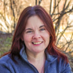 Jess, who has red hair and is wearing a gray zip sweater, is seen from the shoulders up smiling into the camera. In the background are blurred, sunlit bare branches.