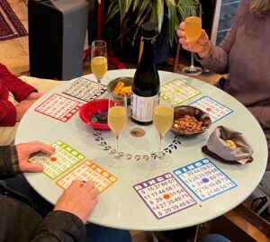 Bingo cards, snacks, and wineglasses on a round table, with people's hands visible on the edges.