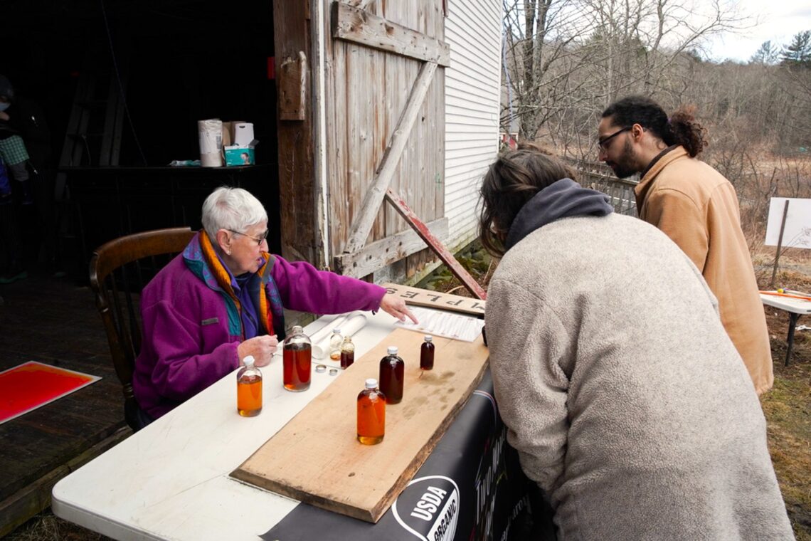 Maple tasting table with a demo of the four grades of maple syrup. Women sitting showing the types of syrup to two tasters.