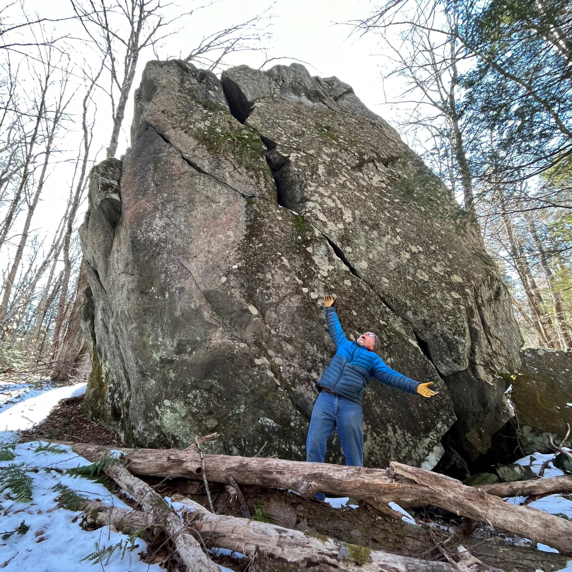 A warmly dressed man poses with his arms flung open, gazing up at a giant snow-ringed boulder.