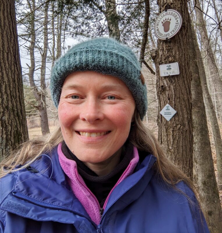 Sarah, warmly dressed, smiles selfie-style into the camera. She is standing in the woods, in front of a tree with Williamsburg Woodland Trails and HLT signs on it.