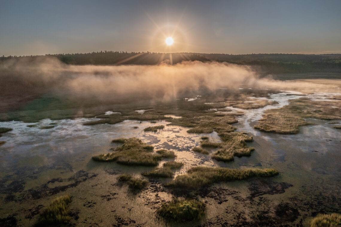 A sunrise over distant hills casts beams of light across a foggy wetland,.