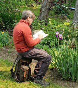 Man sitting on top of a backpack drawing in a nature journal and observing irises.