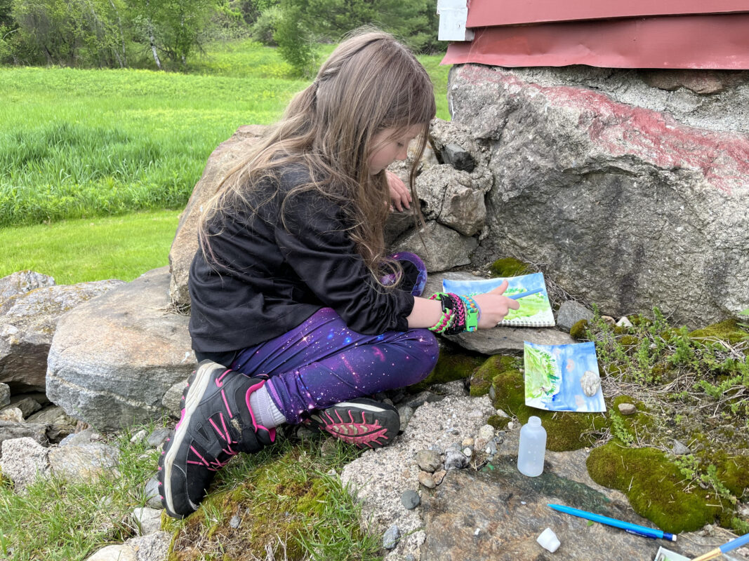 Elementary school aged child with a black shirt and purple pants sits on mossy rocks to the left of the corner of a red barn and the right of a green field. The child is painting two landscapes with bright green and blue watercolors.