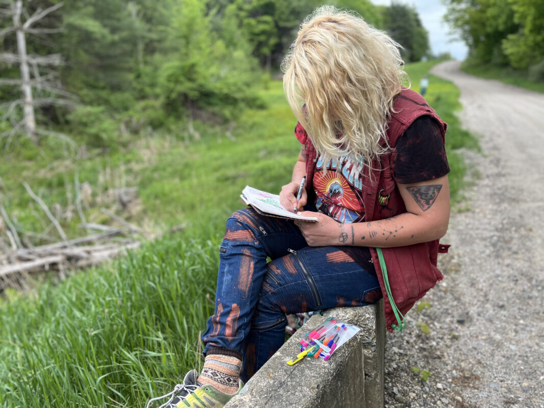 Women sits her leg crossed on the edge of a bridge with green grass and a forest edge to her left and a gravel road to her right. Her blonde wavy hair covers her face as she draws in her nature journal with colored pencils.