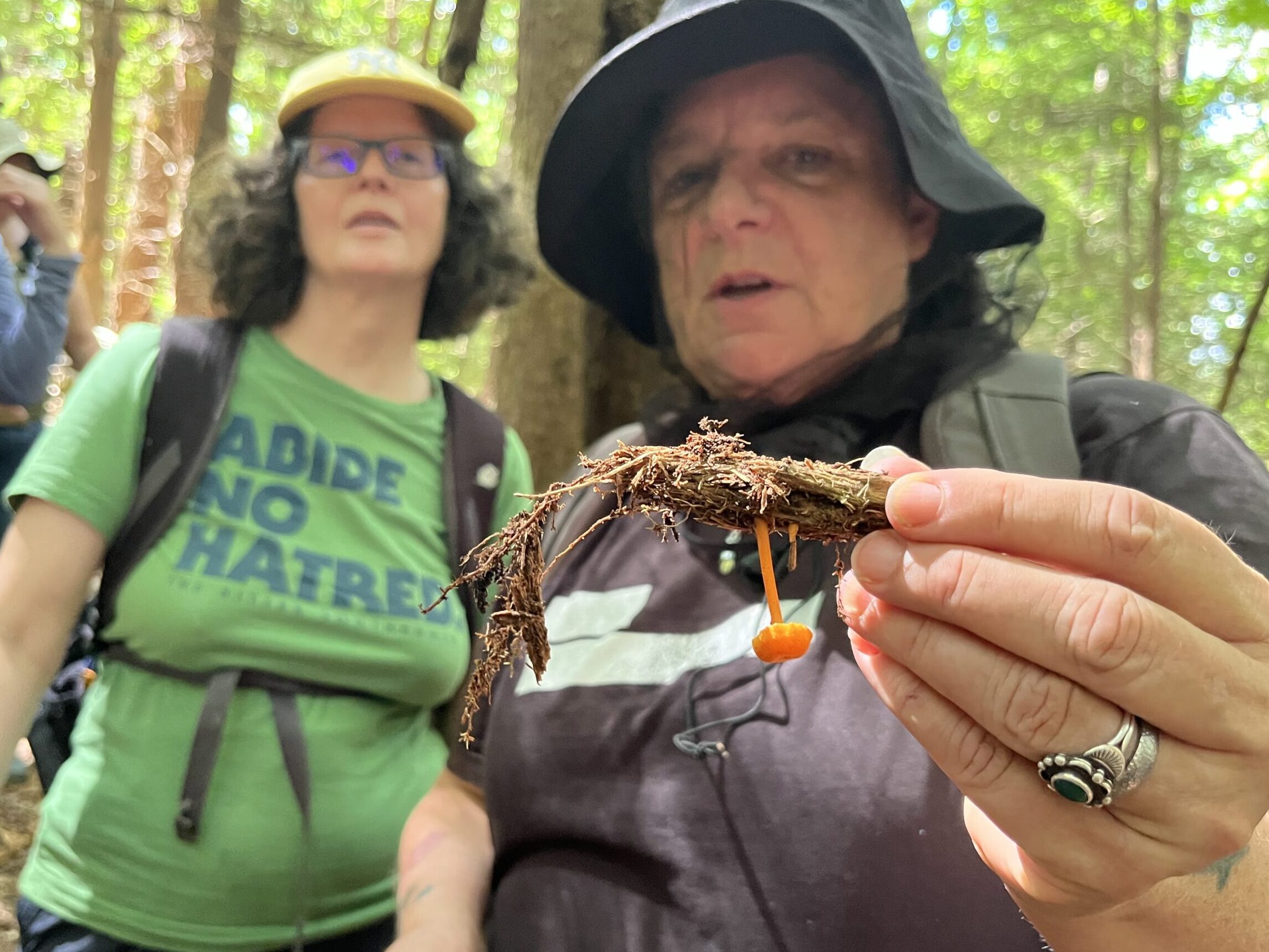 Two women in the frame standing in the woods. One on the right wearing a grey shirt and grey bucket hat is holding bark upside down with a small bright orange Waxy Cap mushroom. 