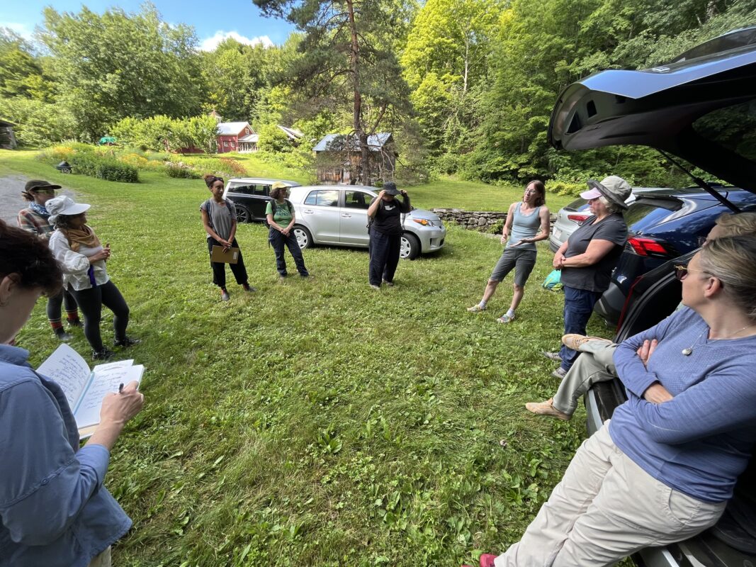 The group of mushroomers stand in a circle next to parked cars in a field along the edge of the forest while Jess Evans talks about the mushroom walk guidelines. 