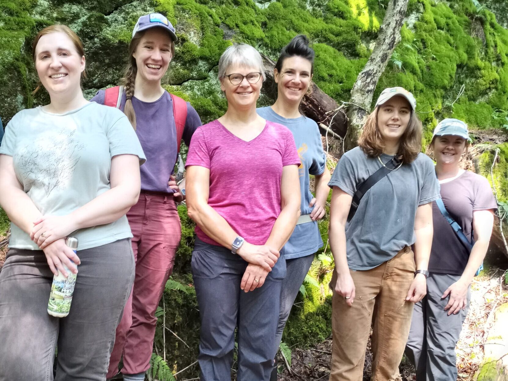 The six members of the HLT staff stand smiling shoulder to shoulder in front of a mossy rock cliff.