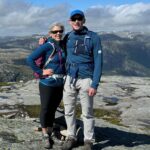 Conan and Cynthia stand arm in arm, wearing hiking clothes and standing in front of a wild mountain landscape.