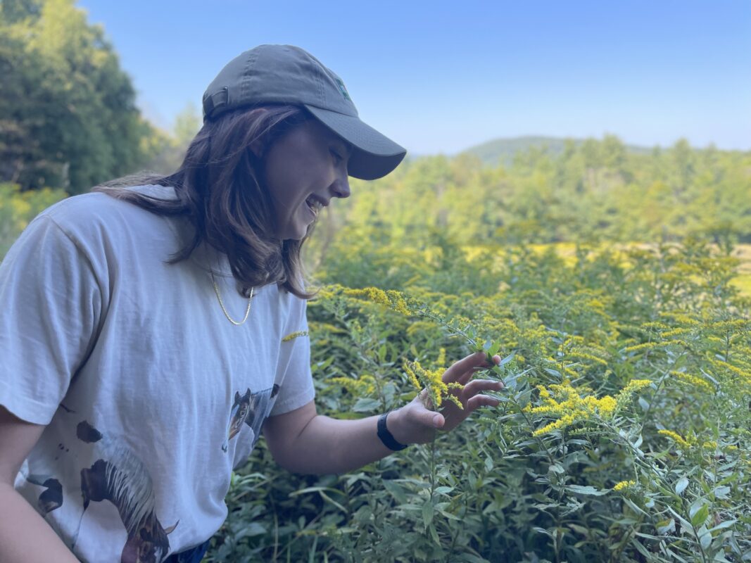 Mariel smiles at a goldenrod flower as she kneels at the edge of a field with a blue sky behind.