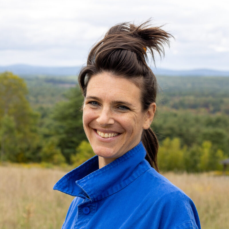 Sara is seen from the shoulders up, smiling towards the camera as she stands in front of a grassy field with a distant view of hills. Her hair is pulled up with a clip and she is wearing a bright blue jacket.