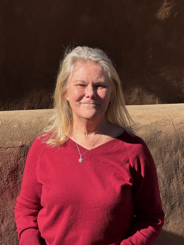 A blonde-haired woman is seen torso up in a portrait-style photo. She is wearing a red shirt and is standing in front of a brown wall in strong sunlight.