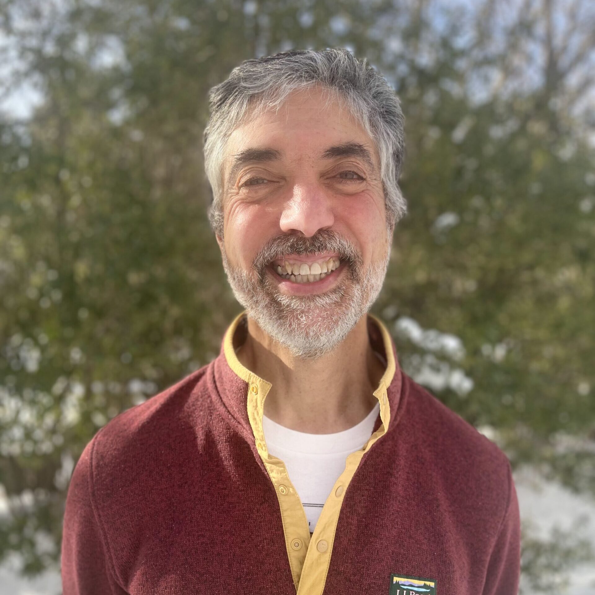 A photo of a smiling man wearing a button-up fleece jacket, with snow and greenery behind him.